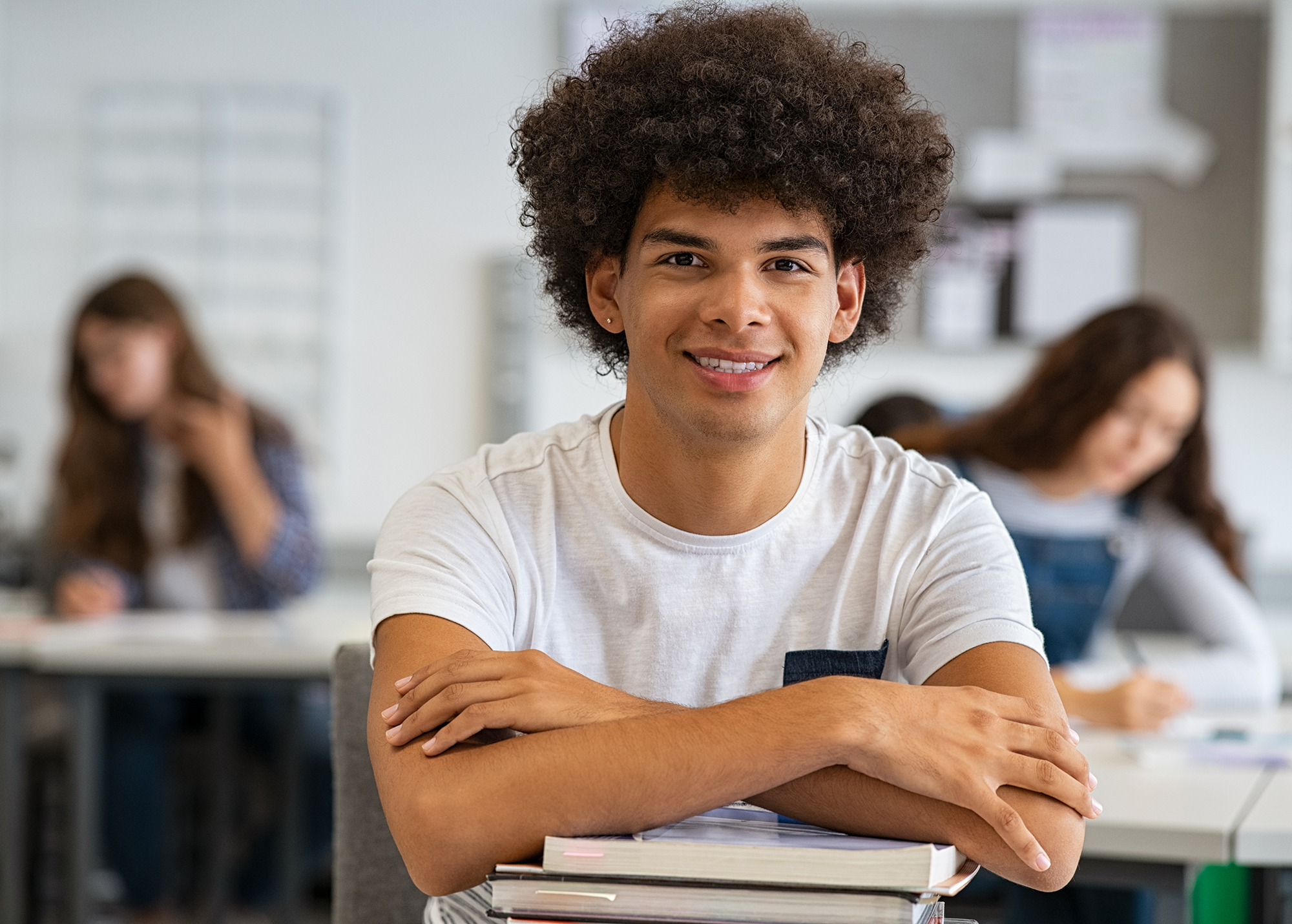 Portrait of happy student in class looking at camera. Smiling young man at college leaning on stack of books with classmates in background.