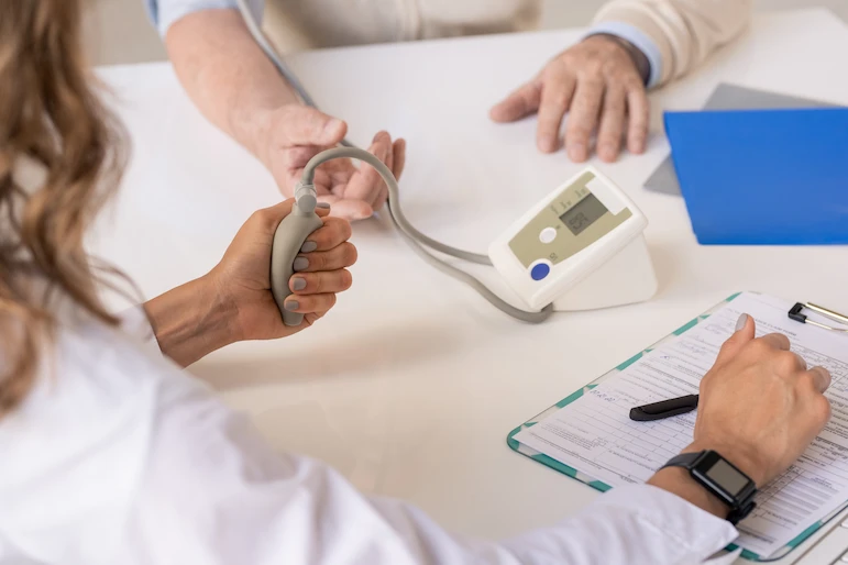A close-up image of a doctor measuring a patient's blood pressure using a digital blood pressure monitor. The doctor's hand is holding the pump while the patient has the cuff on their arm. A clipboard with notes and a pen is on the table.