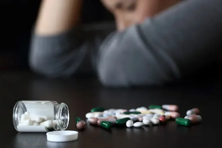 Image showing a person in the background with their head resting on their arm, appearing distressed. In the foreground, a variety of pills and a small open pill bottle are scattered on a dark surface.